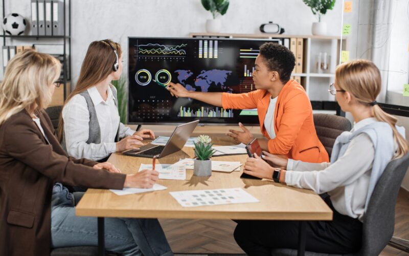 Group of diverse female colleagues looking at computer monitor with worldwide business concept while sitting together at desk. Competent financiers examining graphs, charts and statistics at office.